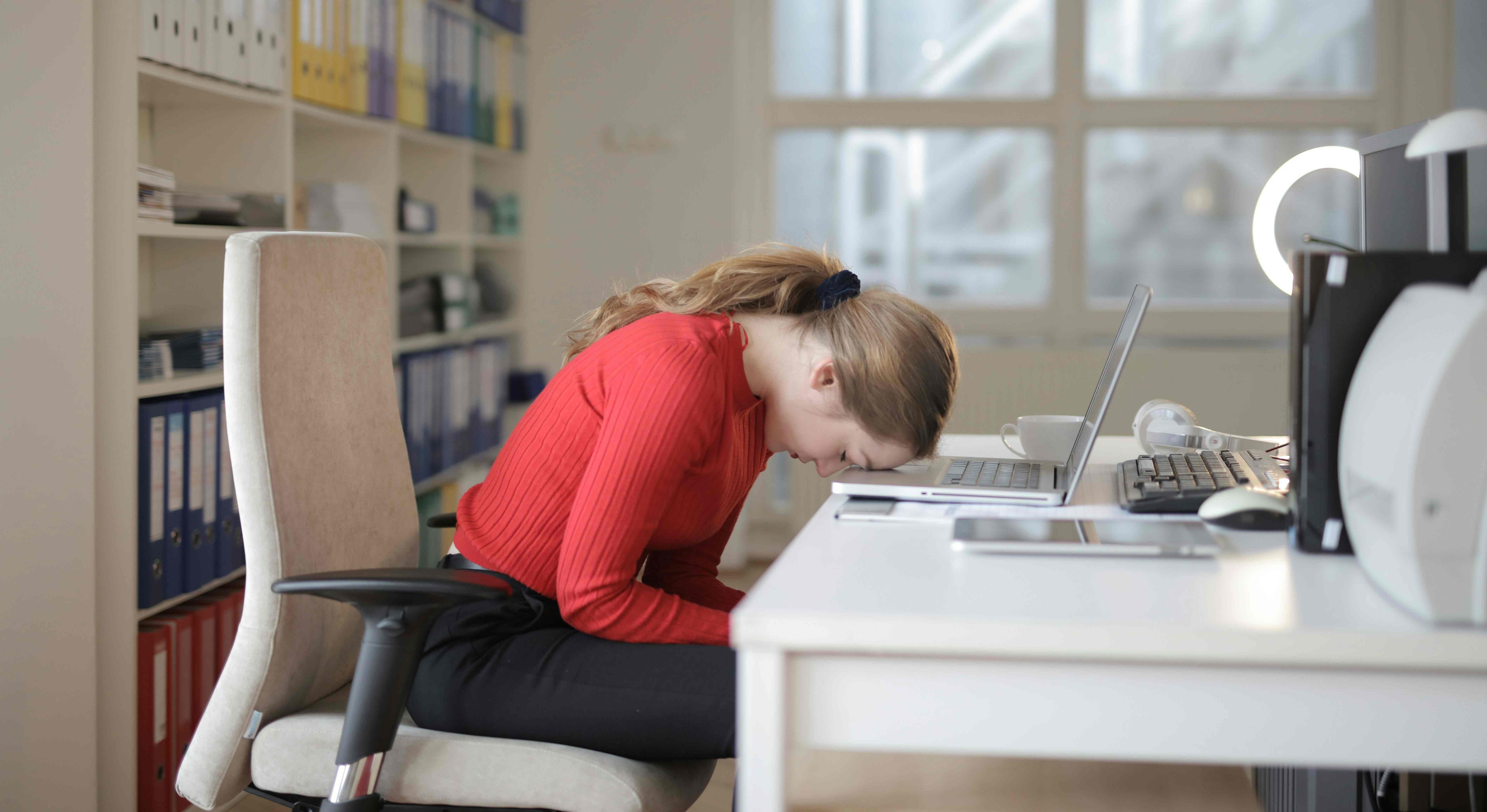 Žena v kanceláři sedí unavená na židli, přičemž má hlavu opřenou o notebook položený na stole.